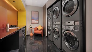 Une buanderie moderne équipée de quatre machines à laver superposées, d’un petit coin salon avec un coussin et d’un comptoir muni d’un distributeur de savon.