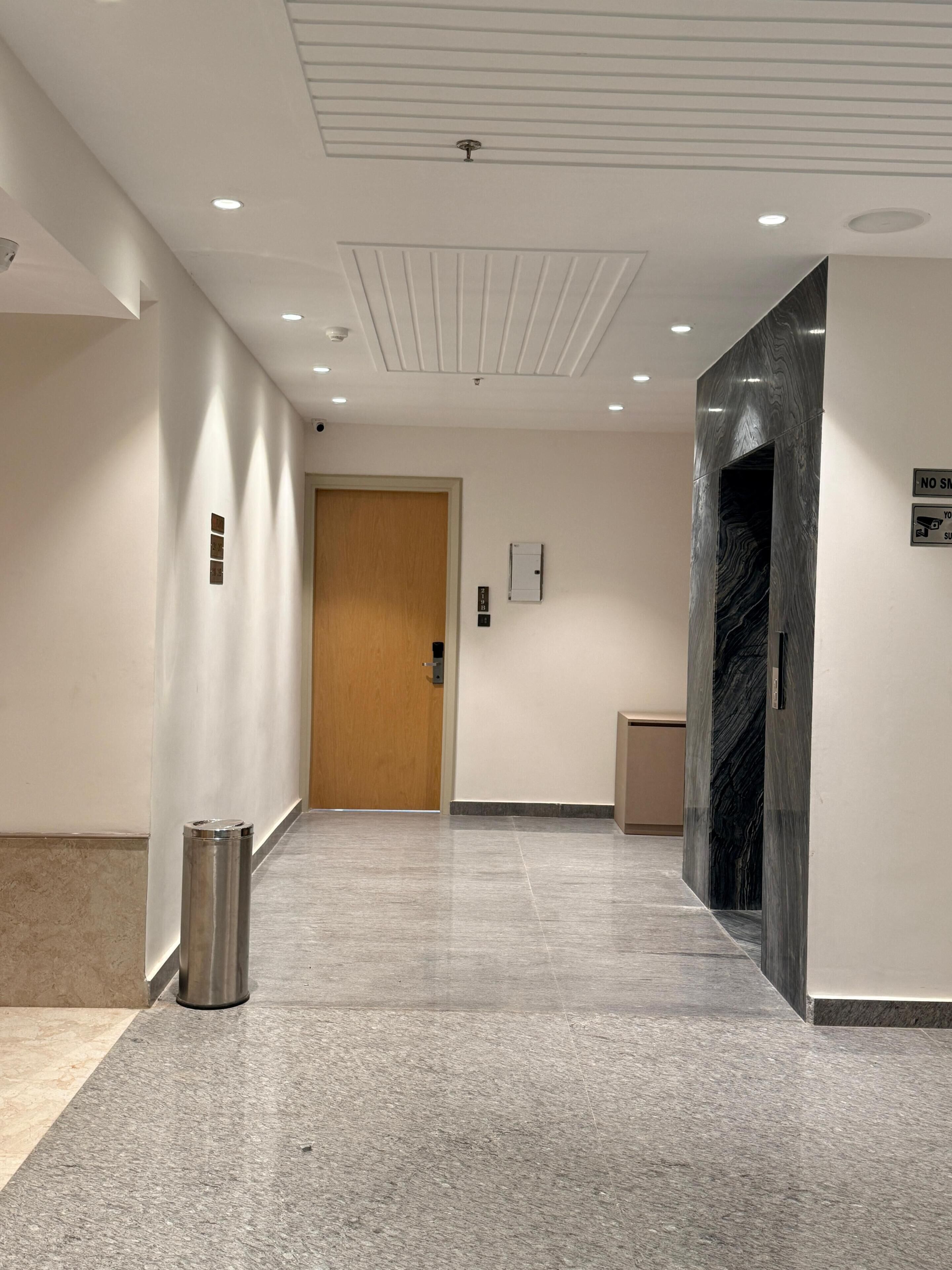 Un pasillo con una puerta de madera, un cesto de basura, un portero automático en la pared y una pared de mármol.