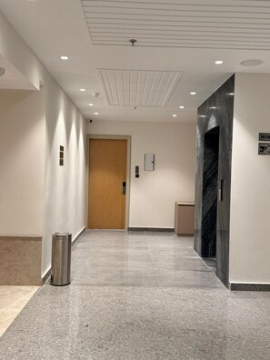 Un pasillo con una puerta de madera, un cesto de basura, un portero automático en la pared y una pared de mármol.