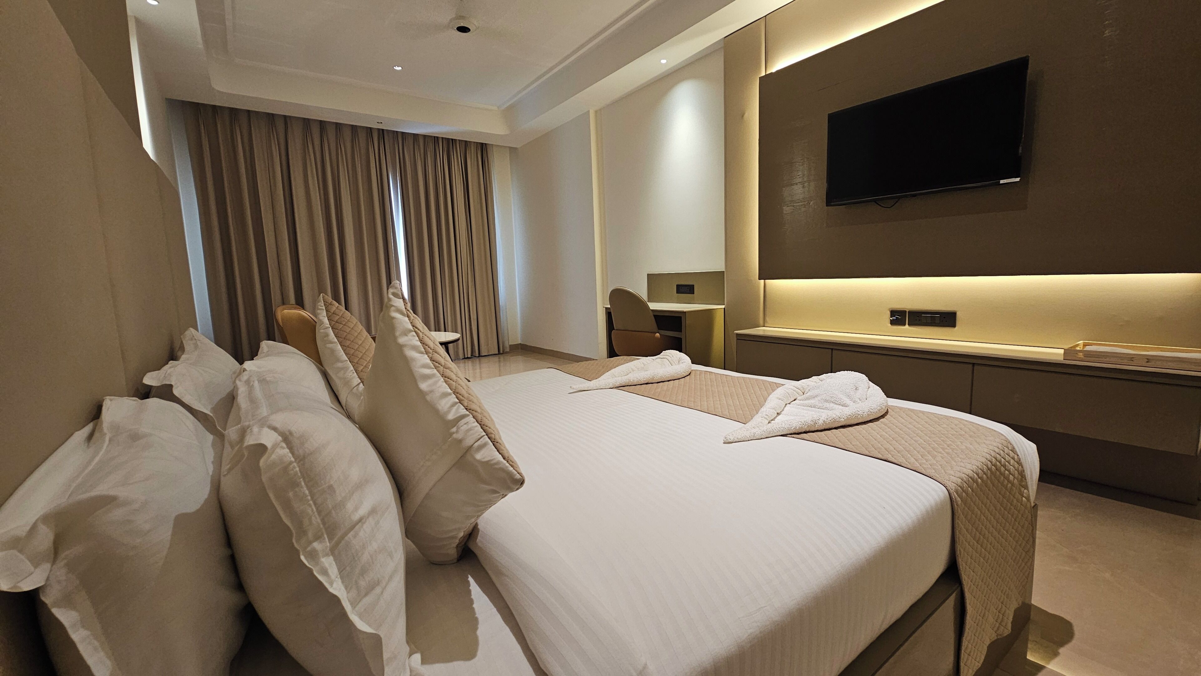 A hotel room with a large bed, a desk, a chair, a TV, and a window with curtains.