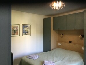 Ein Schlafzimmer mit einem Bett, zwei gerahmten floralen Bildern an der Wand, einer Deckenleuchte und einem Holzregal mit einer kleinen Figur.