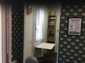Ein kleines Zimmer mit Tisch, Stuhl, einem Fenster mit Jalousien, einer Uhr, einem Regal mit Geschirr und einem Willkommensschild.