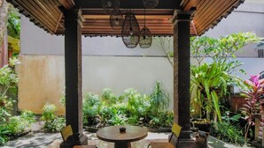 Un patio al aire libre con una mesa redonda y sillas, rodeado de abundante vegetación y una estatua decorativa de un perro.