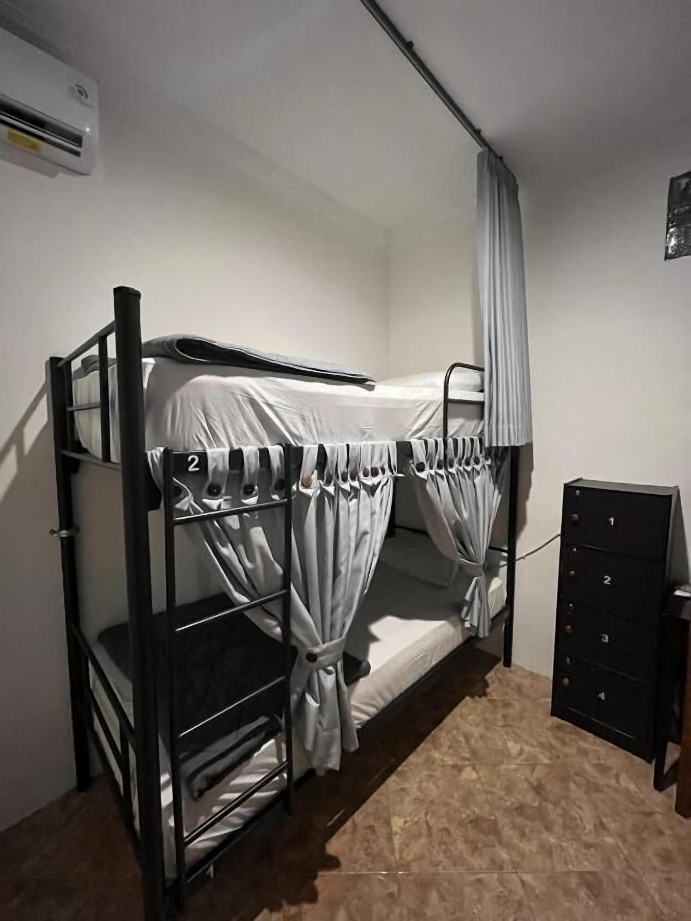 Een stapelbed met een gordijn, een klein kastje en een wandairconditioner.