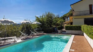 A swimming pool with lounge chairs and umbrellas, surrounded by greenery and a building. - "The Infinity" Apartment of Sibillini Montains (Montefortino)