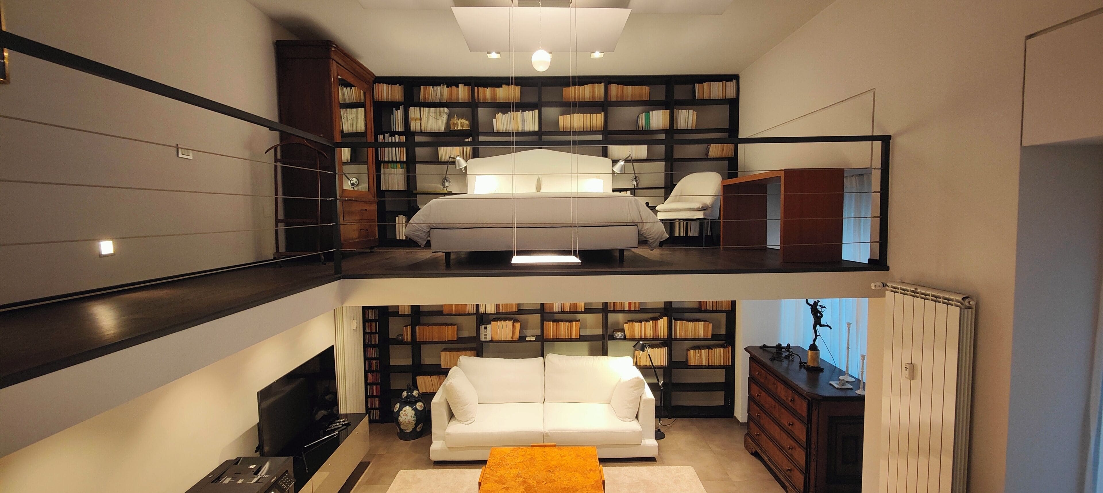 A modern loft-style apartment with a loft bed, a white sofa, a fireplace, and a large bookshelf.
