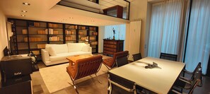 Ein modernes Wohnzimmer mit einem weißen Sofa, einem Esstisch mit Stühlen, einem Bücherregal und einer Heizung.