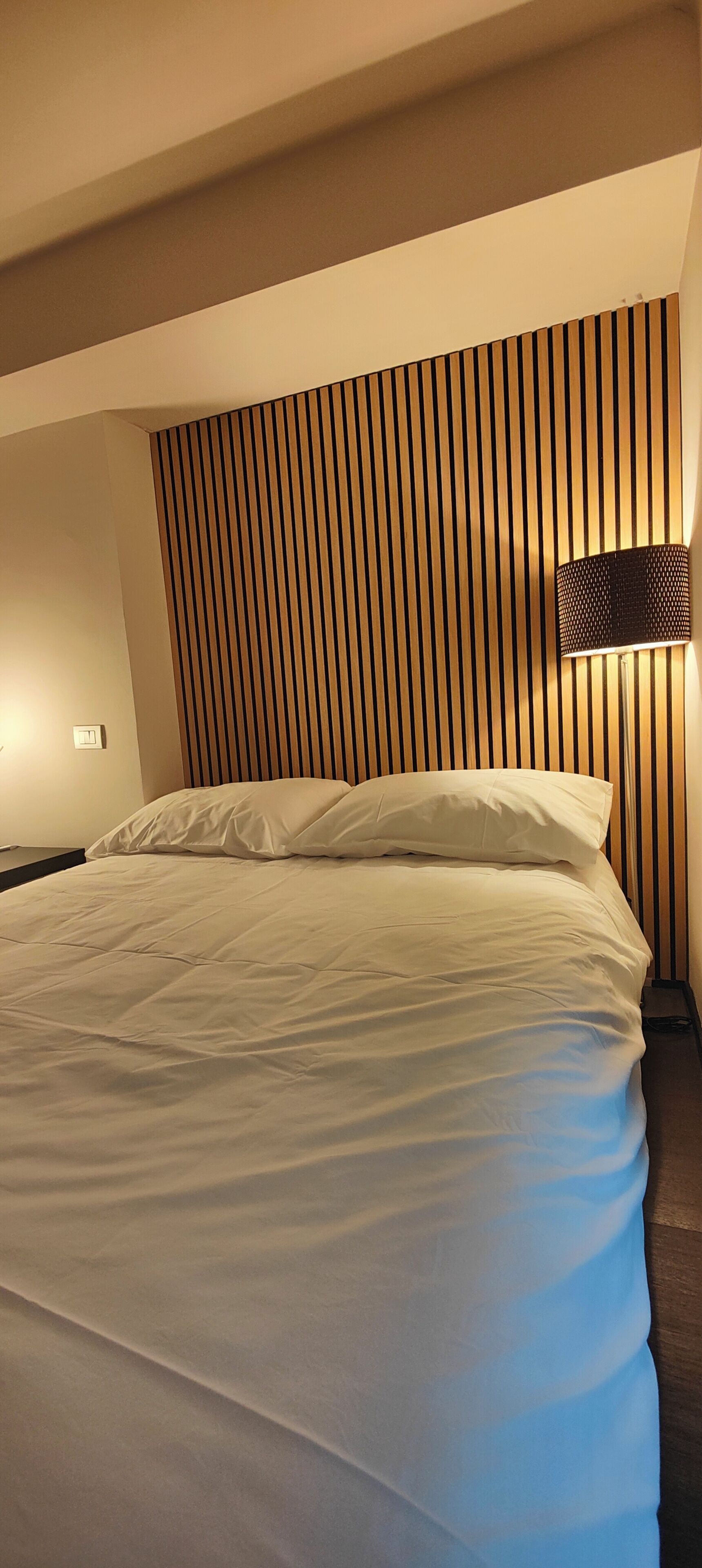 A hotel room with a bed, a wall-mounted lamp, and a wooden slat headboard.