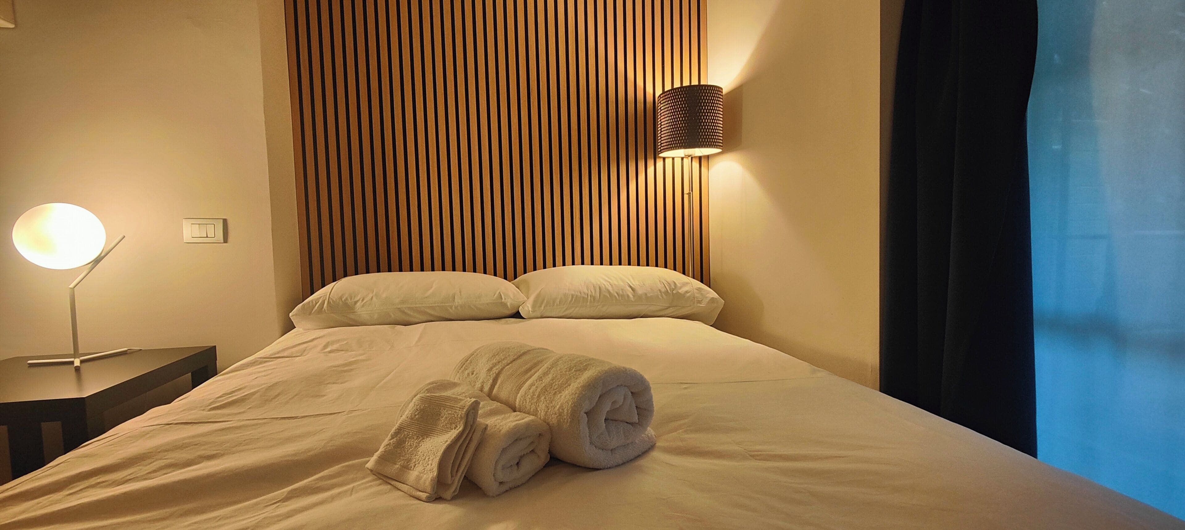 A neatly made bed with pillows and rolled towels, a bedside lamp, and a wall with vertical wooden slats.