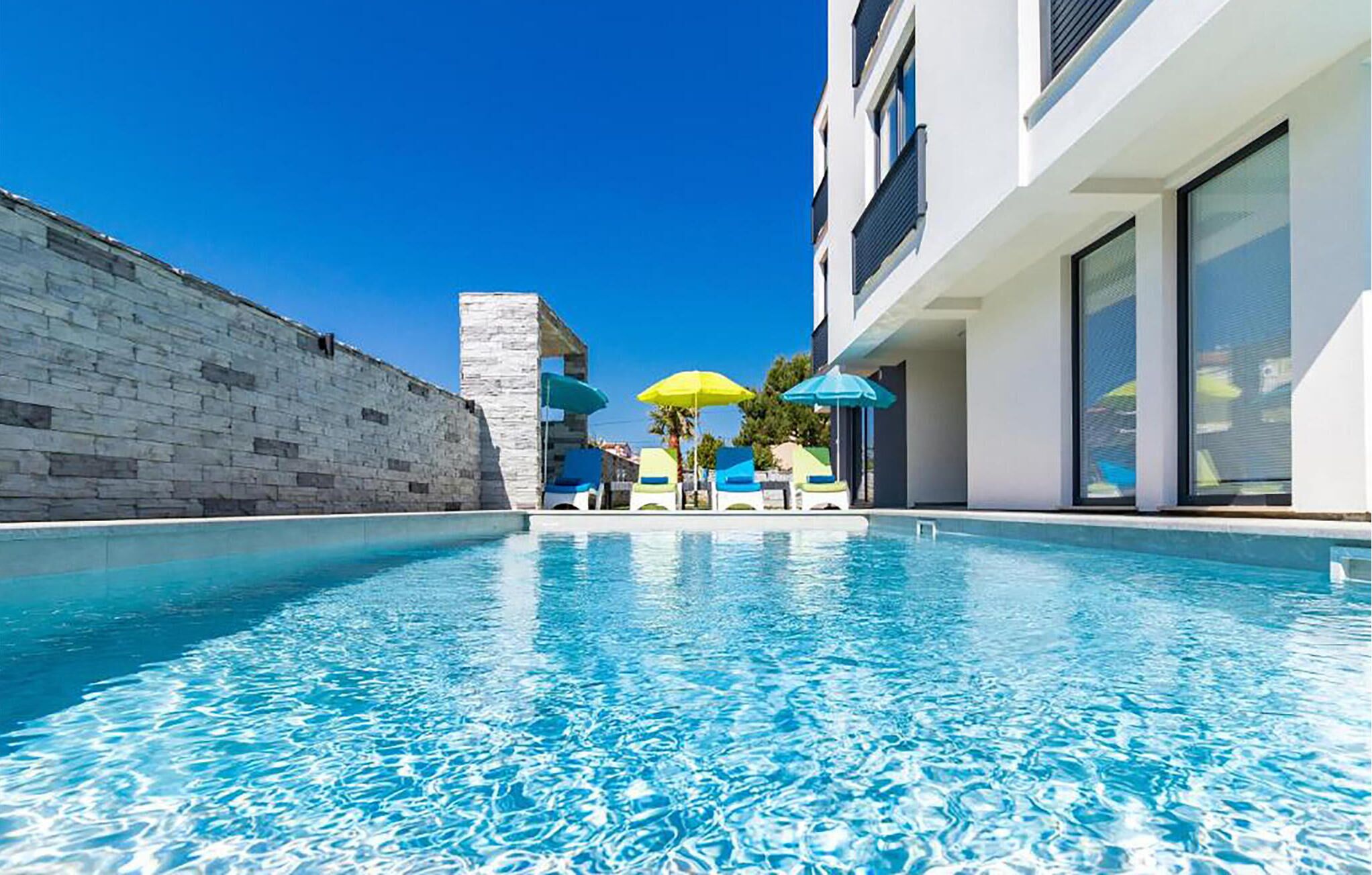 A swimming pool with lounge chairs and umbrellas, a stone wall, and a modern building with large windows.