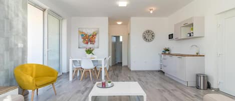 Una sala moderna con un sofá, un sillón amarillo, un comedor con mesa y sillas, una cocina con armarios blancos y un reloj, y un pasillo que conduce a otra habitación.