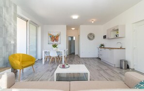 Ein modernes Wohnzimmer mit einer Couch, einem gelben Sessel, einem Essbereich mit Tisch und Stühlen, einer Küche mit weißen Schränken und einer Uhr sowie einem Flur, der in einen anderen Raum führt.
