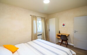 Ein Schlafzimmer mit einem Bett, einem Schreibtisch, einem Stuhl, einer Tür und einem Fenster mit Vorhängen.