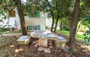 An outdoor stone table with benches, surrounded by trees and a building with green shutters. - Stunning home in Krnica (Krnica)