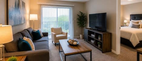 A modern living room with a sofa, armchair, coffee table, TV, and a bed in the adjacent room.