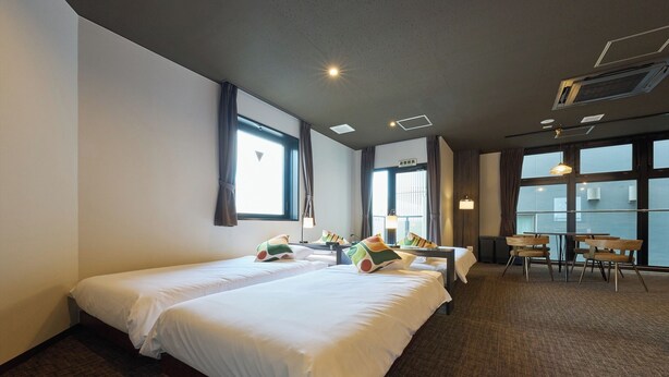 Habitación de hotel con dos camas, una mesita, sillas y ventanas amplias.