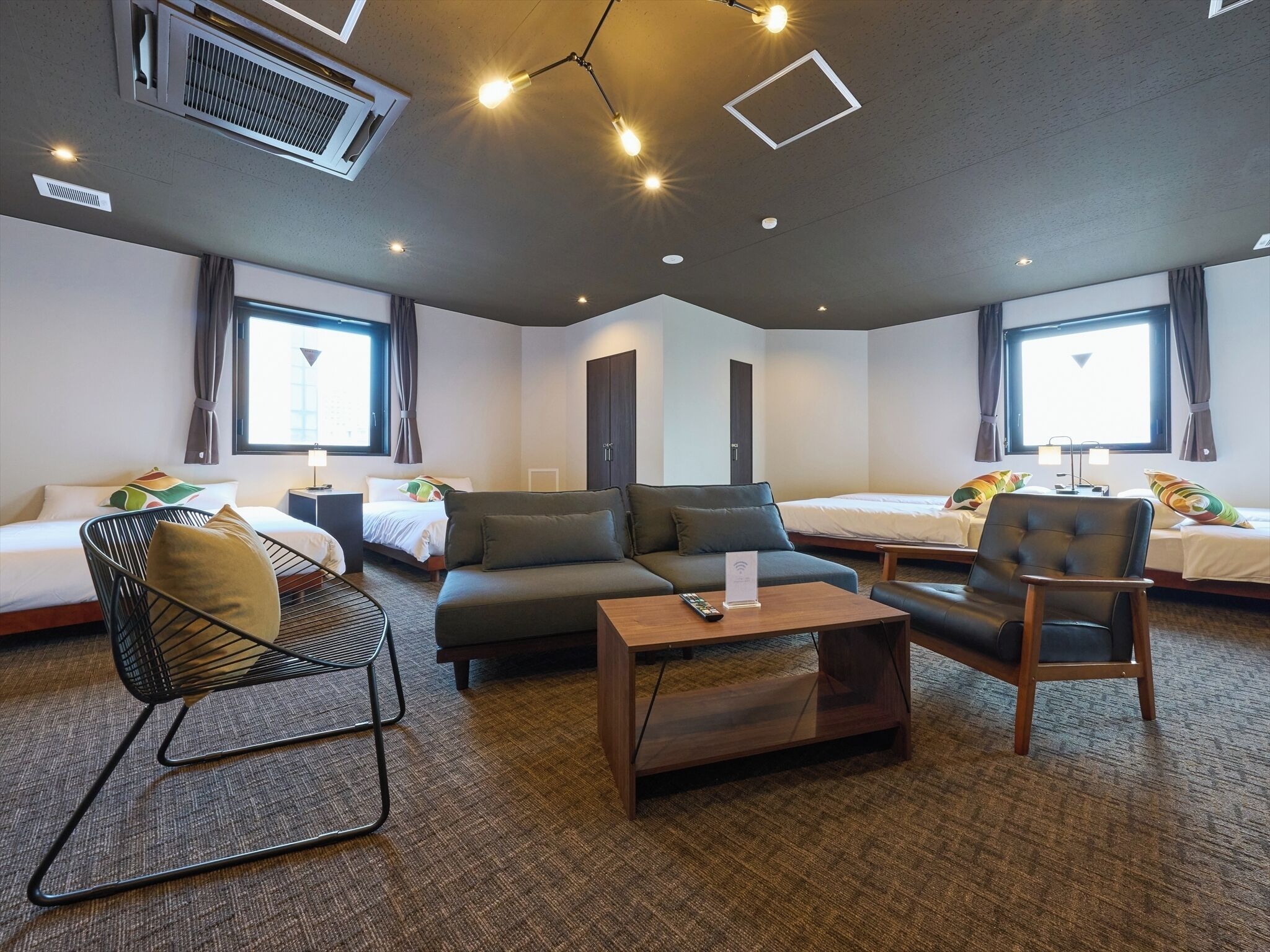 A modern hotel room with a sofa, armchair, and a small table. There are two beds with headboards and a window with curtains.