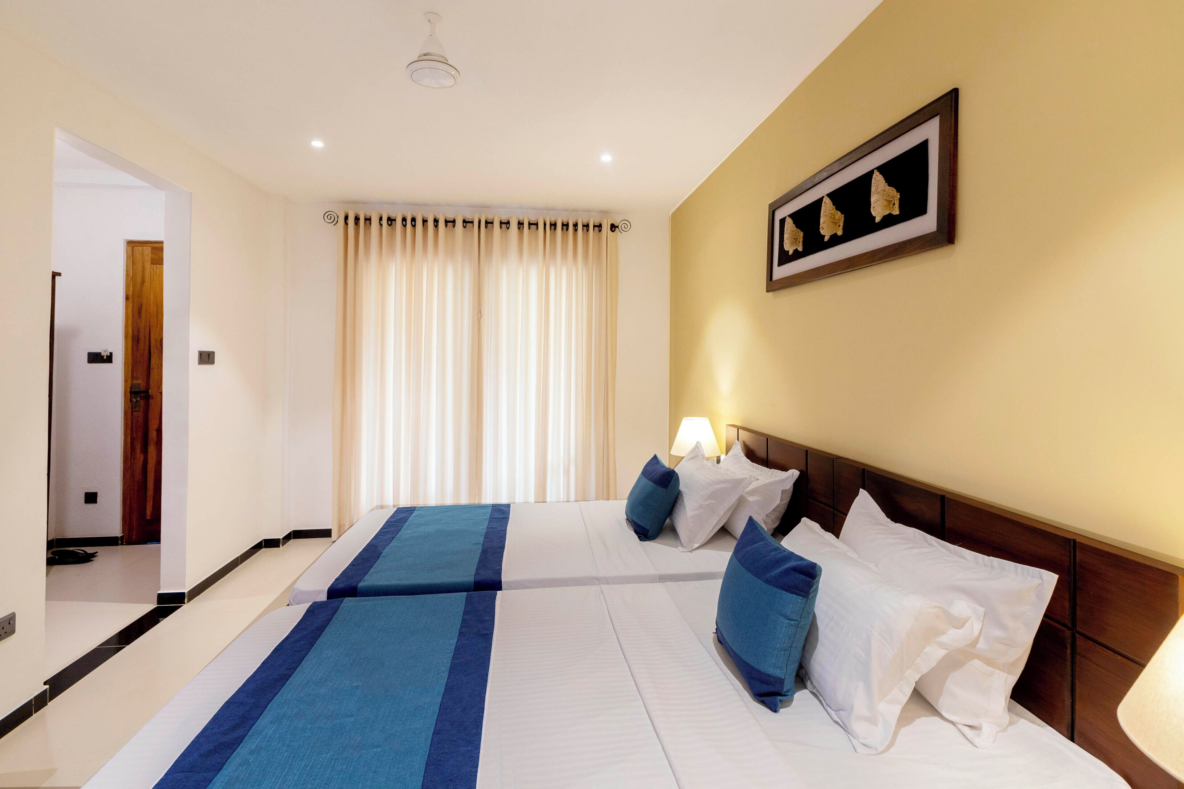 A hotel room with two beds, a headboard, a nightstand, a lamp, a framed picture on the wall, and a window with curtains.