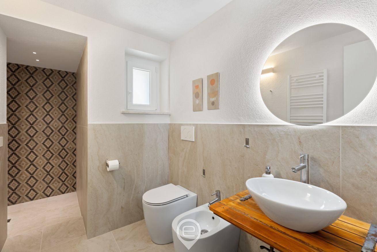A modern bathroom with a large oval mirror, a wooden vanity, a white sink, and a toilet.