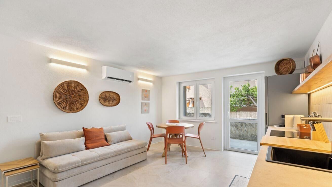 A modern living space with a sofa, dining table, and chairs, a kitchen area, and a large window providing natural light.