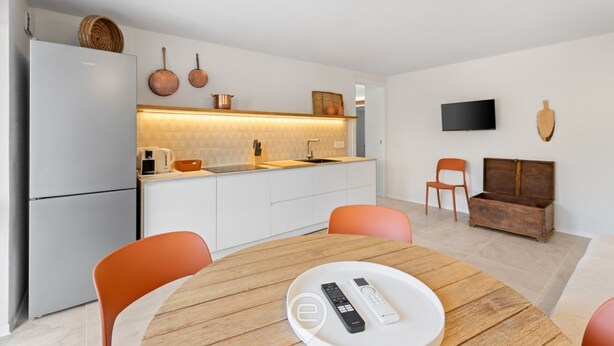 Una cocina moderna con armarios blancos, horno integrado y comedor con mesa de madera y sillas naranjas.