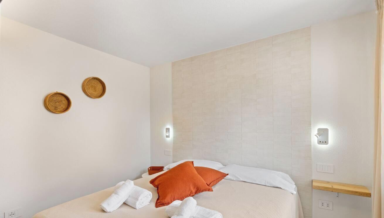 A hotel room with a bed, two wall-mounted light fixtures, a wooden shelf, and two wall decorations.