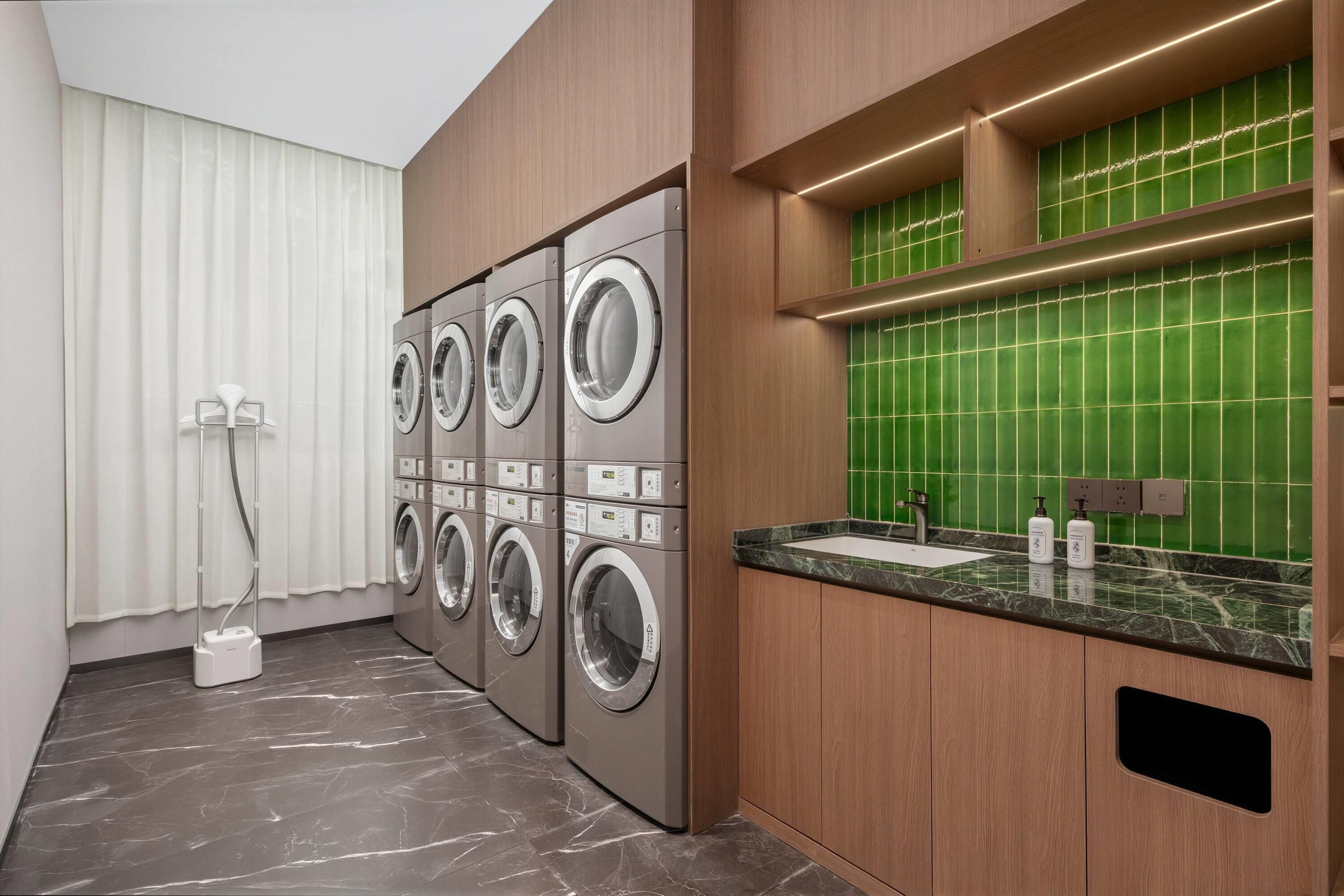 A modern laundry room with four stacked front-loading washing machines, a sink with a marble countertop, and a green-tiled backsplash.