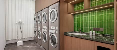 A modern laundry room with four stacked front-loading washing machines, a sink with a marble countertop, and a green-tiled backsplash.