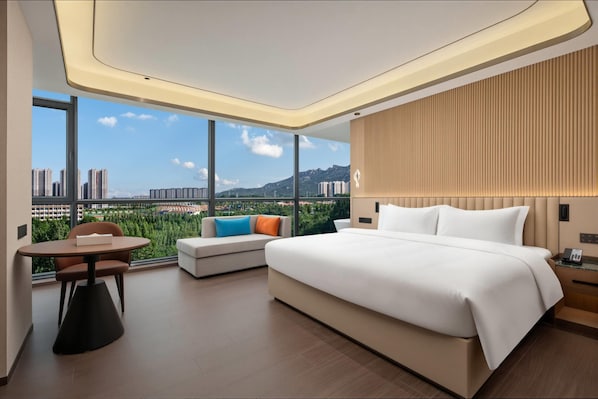 Quarto de hotel moderno com uma cama grande, área de estar com uma mesa e vista para a paisagem urbana através de amplas janelas.