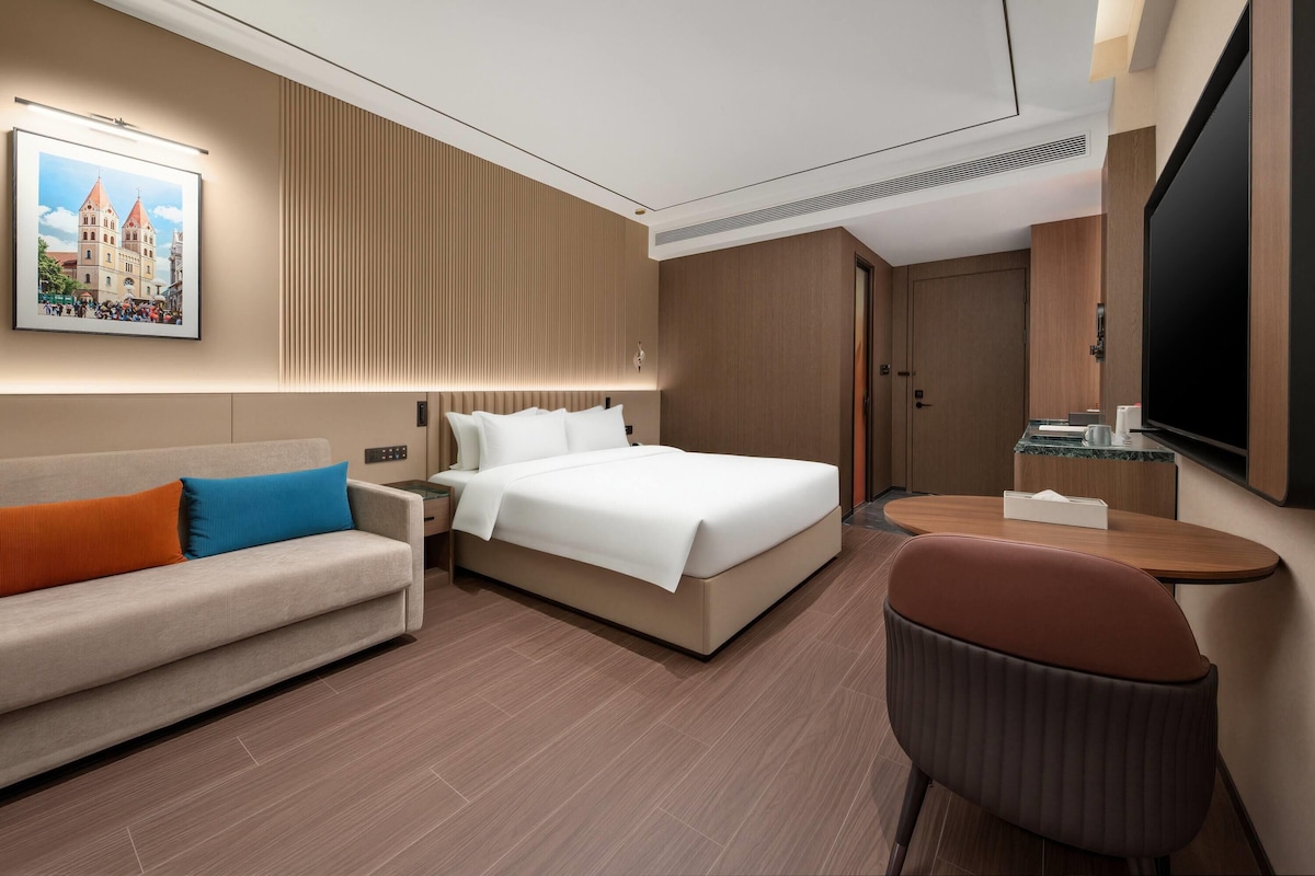 Quarto de hotel com cama, sofá, escrivaninha, poltrona, televisão e um quadro na parede.