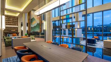 A modern hotel lobby with a large conference table, seating, a reception desk, and a view of a swimming pool.