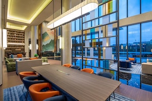 A modern hotel lobby with a large conference table, seating, a reception desk, and a view of a swimming pool.