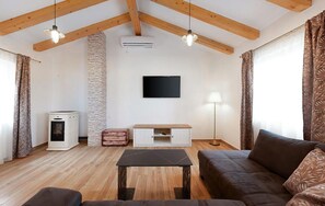 A living room with a wooden ceiling, a flat-screen TV mounted on the wall, a white entertainment unit, a brown sofa, a small table, and a lamp. - Stunning home in Jurdani (Jurdani)