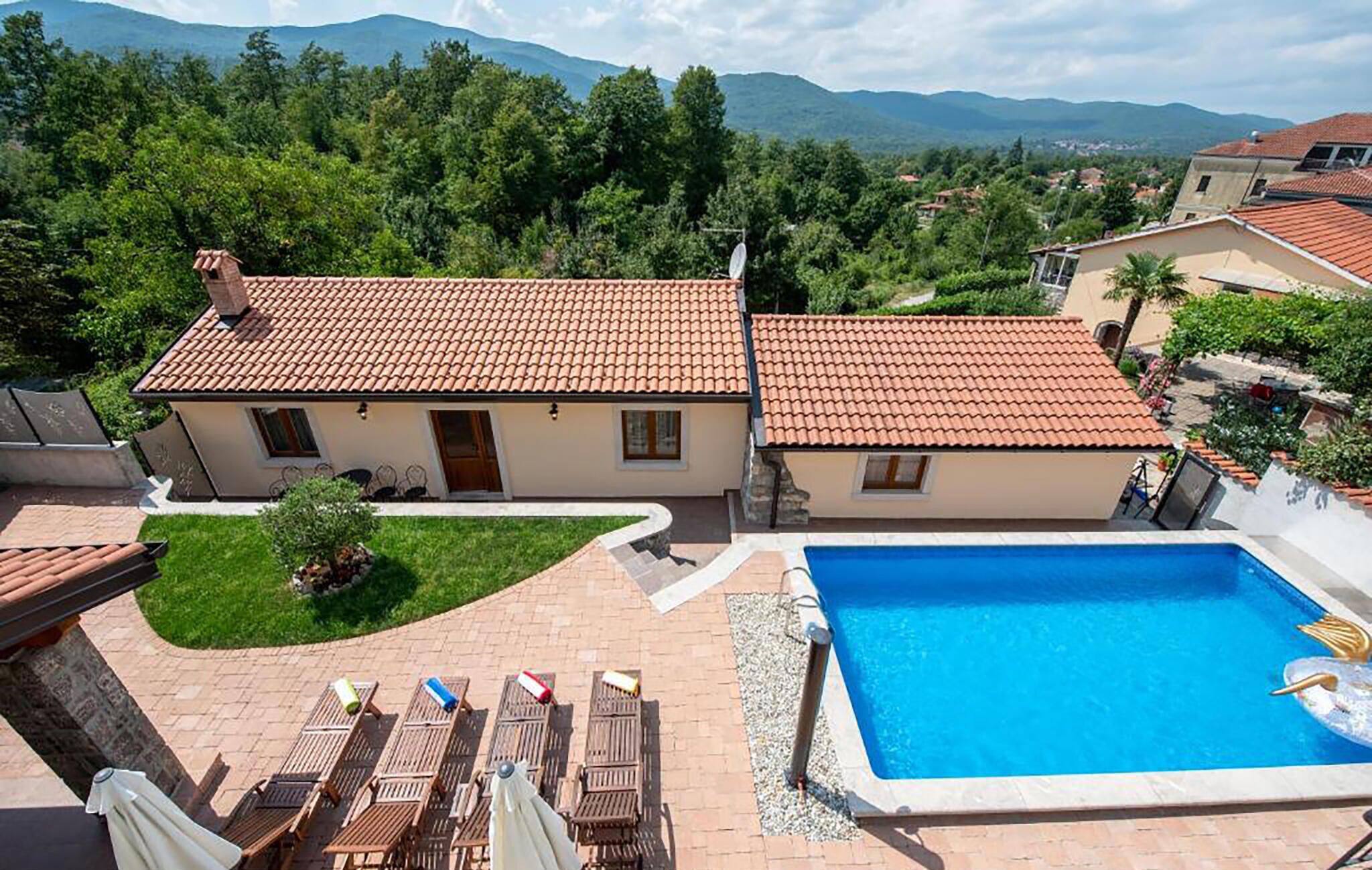 A two-story house with a swimming pool, surrounded by greenery and mountains in the background.