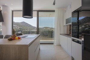 A modern kitchen with a large island, a built-in oven, and a view of a hilly landscape. - Gaia Villas - With 2 Private Pools (Bali)