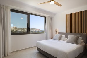 A modern bedroom with a large window, a bed with pillows, a wooden panel wall, and a ceiling fan. - Gaia Villas - With 2 Private Pools (Bali)