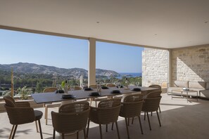 A dining area with a table set for six, wicker chairs, a stone wall, and a view of the sea and hills. - Gaia Villas - With 2 Private Pools (Bali)