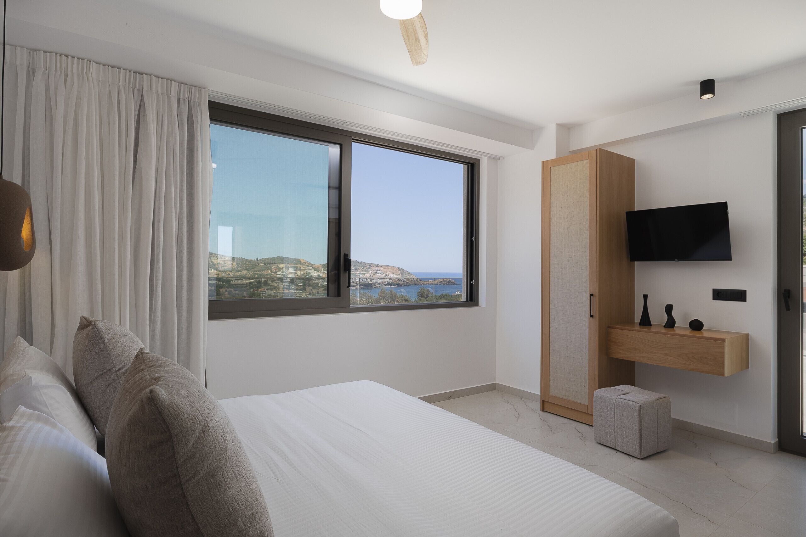 Ein modernes Schlafzimmer mit einem großen Bett, einem an der Wand montierten Fernseher und Meerblick durch ein großes Fenster.