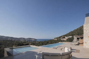 Ein Dachpool mit zwei Bereichen, umgeben von Liegestühlen und Sonnenschirmen, mit Blick auf eine Küstenlandschaft mit Hügeln und blauem Himmel.