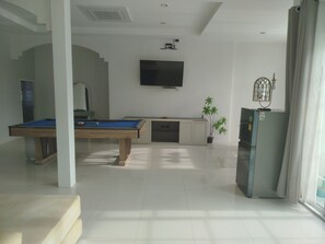 A room with a pool table, a flat-screen TV mounted on the wall, a refrigerator, and a potted plant.