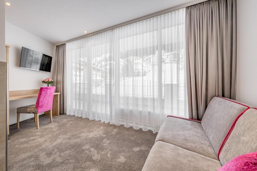 Een hotelkamer met een groot raam, een flatscreen televisie aan de muur, een houten bureau, een roze stoel en een beige bank.