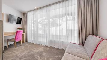 Een hotelkamer met een groot raam, een flatscreen televisie aan de muur, een houten bureau, een roze stoel en een beige bank.