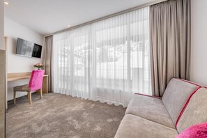 Habitación de hotel con un ventanal grande, un televisor de pantalla plana montado en la pared, un escritorio de madera, una silla rosa y un sofá color beige.
