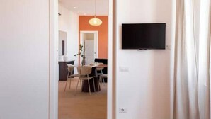 Una habitación con un televisor de pantalla plana montado en la pared, un comedor con sillas y una mesa, y una puerta que da a otra habitación.