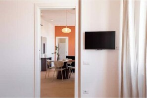 Ein Zimmer mit einem an der Wand befestigten Flachbildfernseher, einem Essbereich mit Stühlen und einem Tisch sowie einer Tür zu einem anderen Raum.