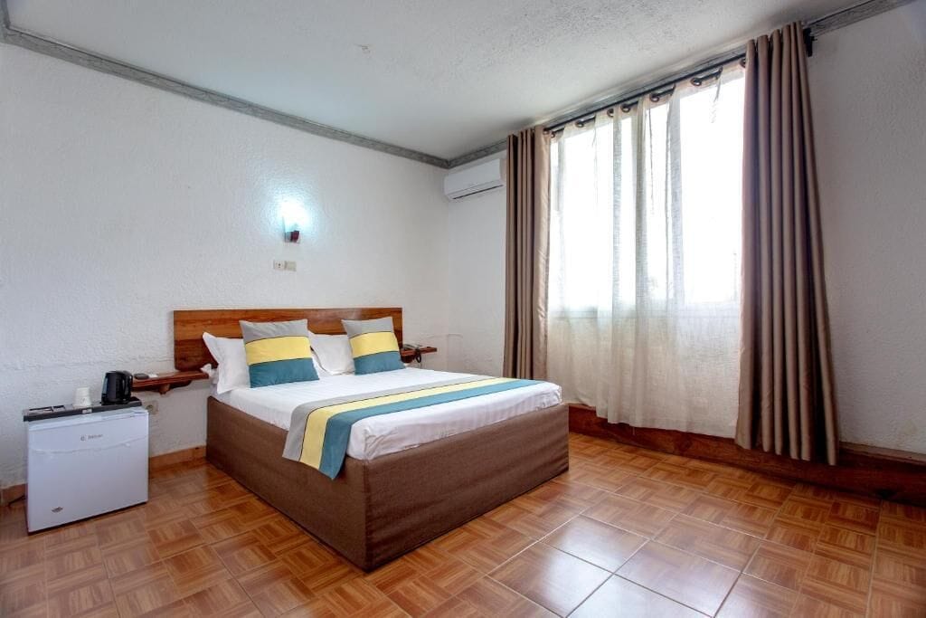 Quarto de hotel com cama, mesa de cabeceira e um pequeno eletrodoméstico branco.