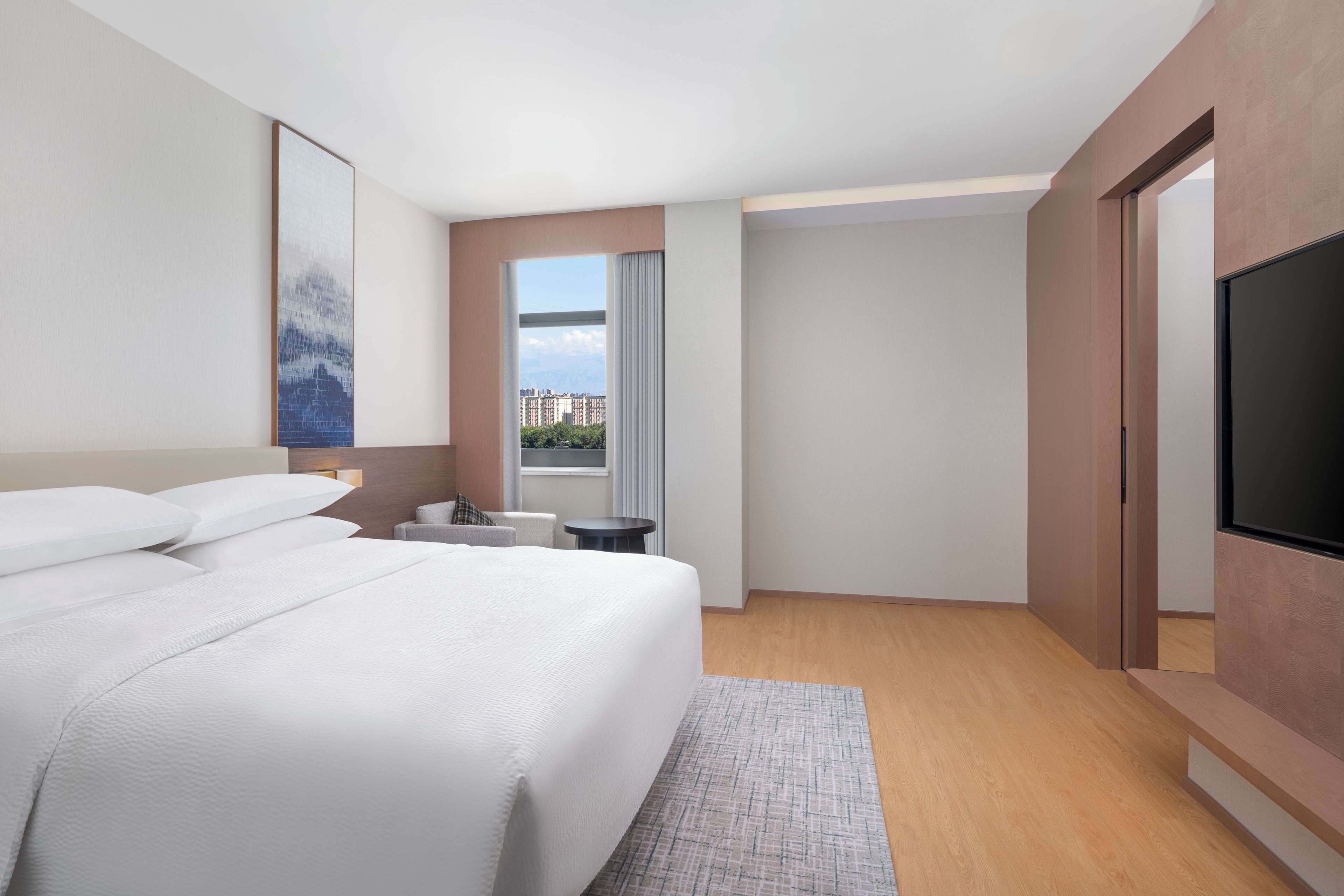 A modern hotel room with a large bed, a flat-screen TV, a window with a city view, and a small table.