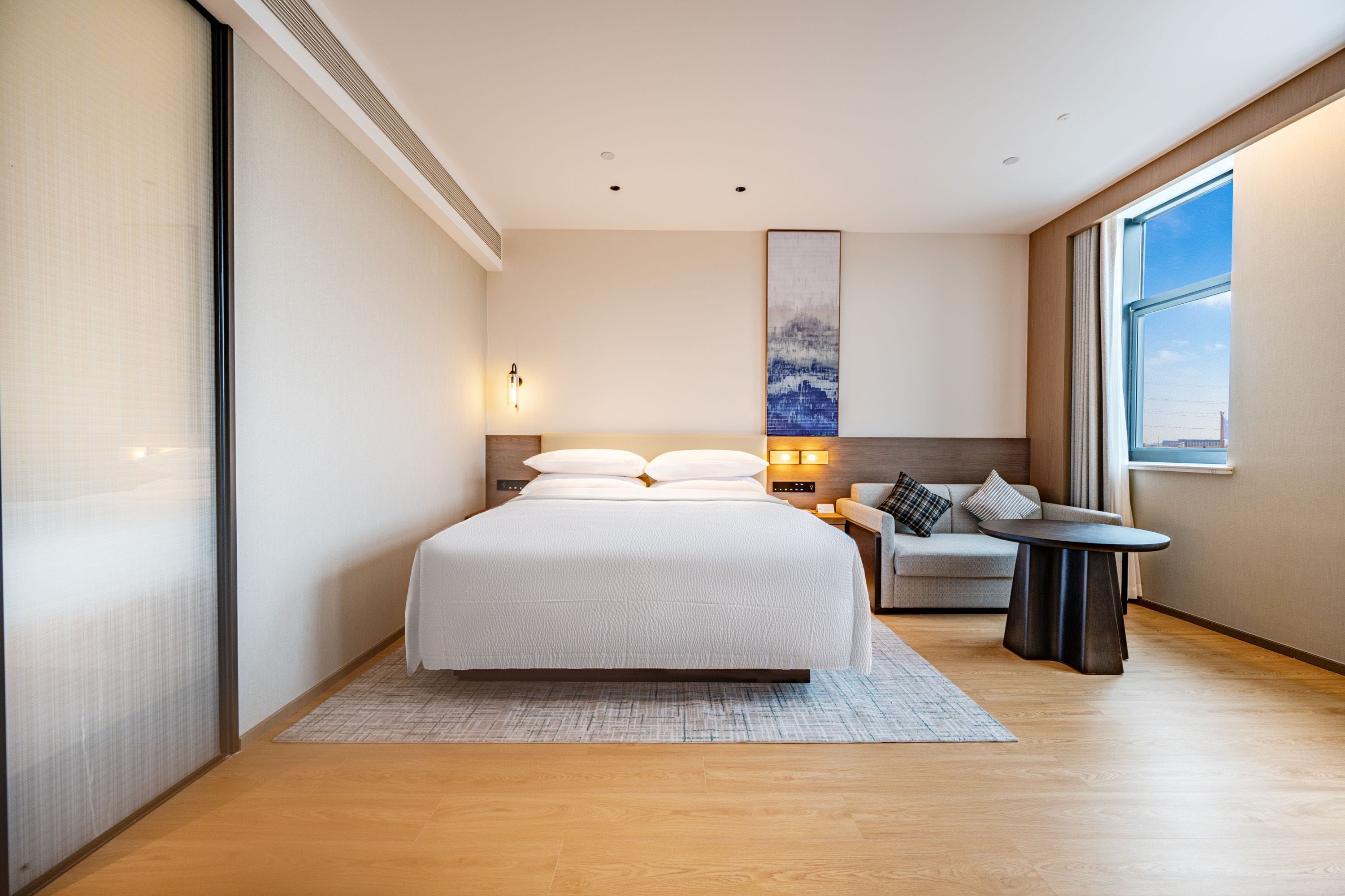 A modern hotel room with a large bed, a seating area with a table, and a large window providing a view.