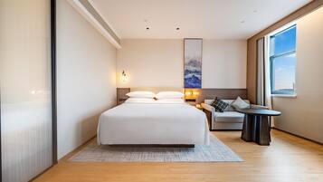 A modern hotel room with a large bed, a seating area with a table, and a large window providing a view.