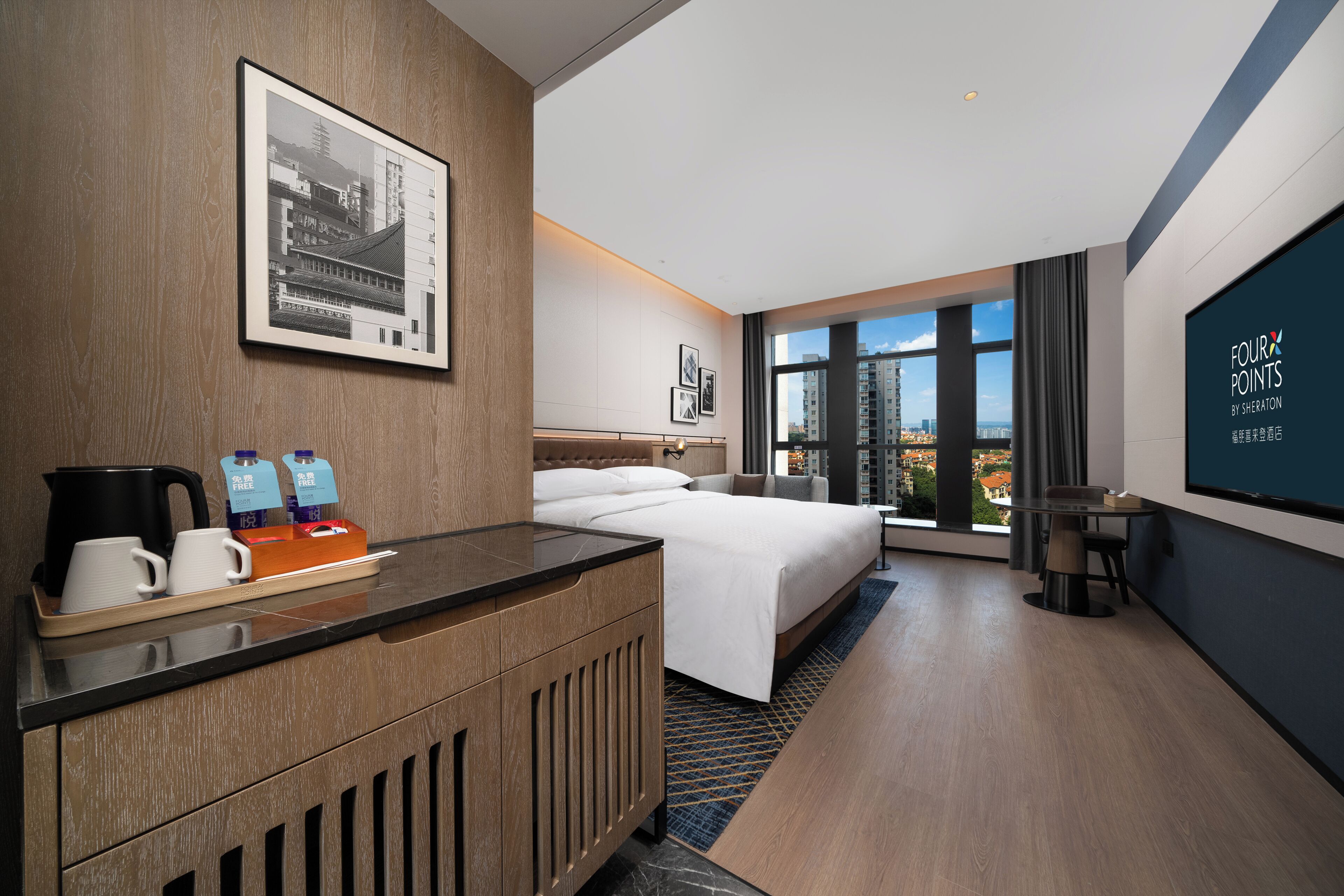 Ein Hotelzimmer mit einem Bett, einem Schreibtisch mit Wasserkocher, Tassen und Tablett, einem großen Fenster mit Blick auf die Stadt und einem gerahmten Bild an der Wand.